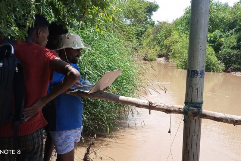 Three men gather round a laptop in a shaded spot next to a water monitoring station located in shallow water, with lush greenery around them